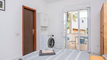 Un dormitorio con cama, lavadora, puerta de madera y puerta corrediza de vidrio que da a un área exterior con sillas y una mesa.