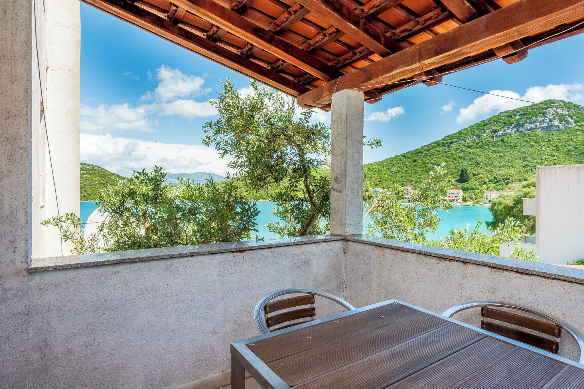 Ein Balkon mit einem Holztisch und Stühlen, mit Blick auf eine Berglandschaft und ein Gewässer.
