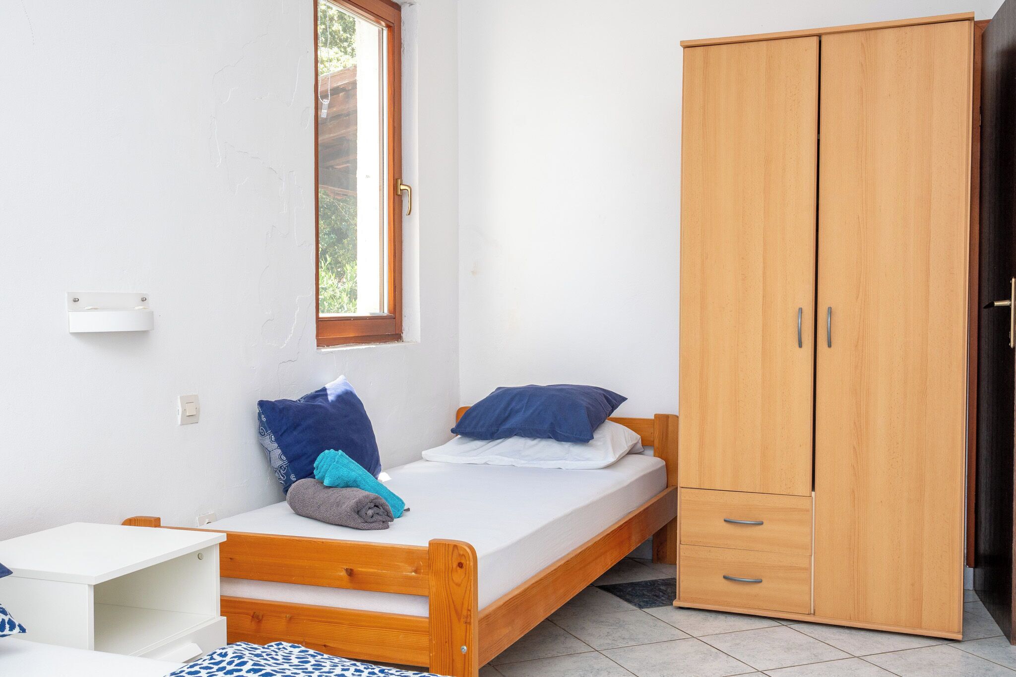 Een enkel bed met houten frame, een klein nachtkastje, een kledingkast en een raam met uitzicht op groen.