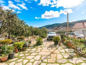 A stone pathway leading to a white car parked in a garden with potted plants and a mountainous backdrop. - Apartments Mirkovic (Marina)