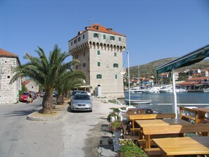 A coastal town with a prominent stone tower, a marina with boats, and outdoor seating by the water. - Apartments Mirkovic (Marina)