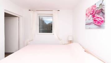 A bedroom with a bed, a window with curtains, a bedside table with a lamp, and a wall-mounted picture.