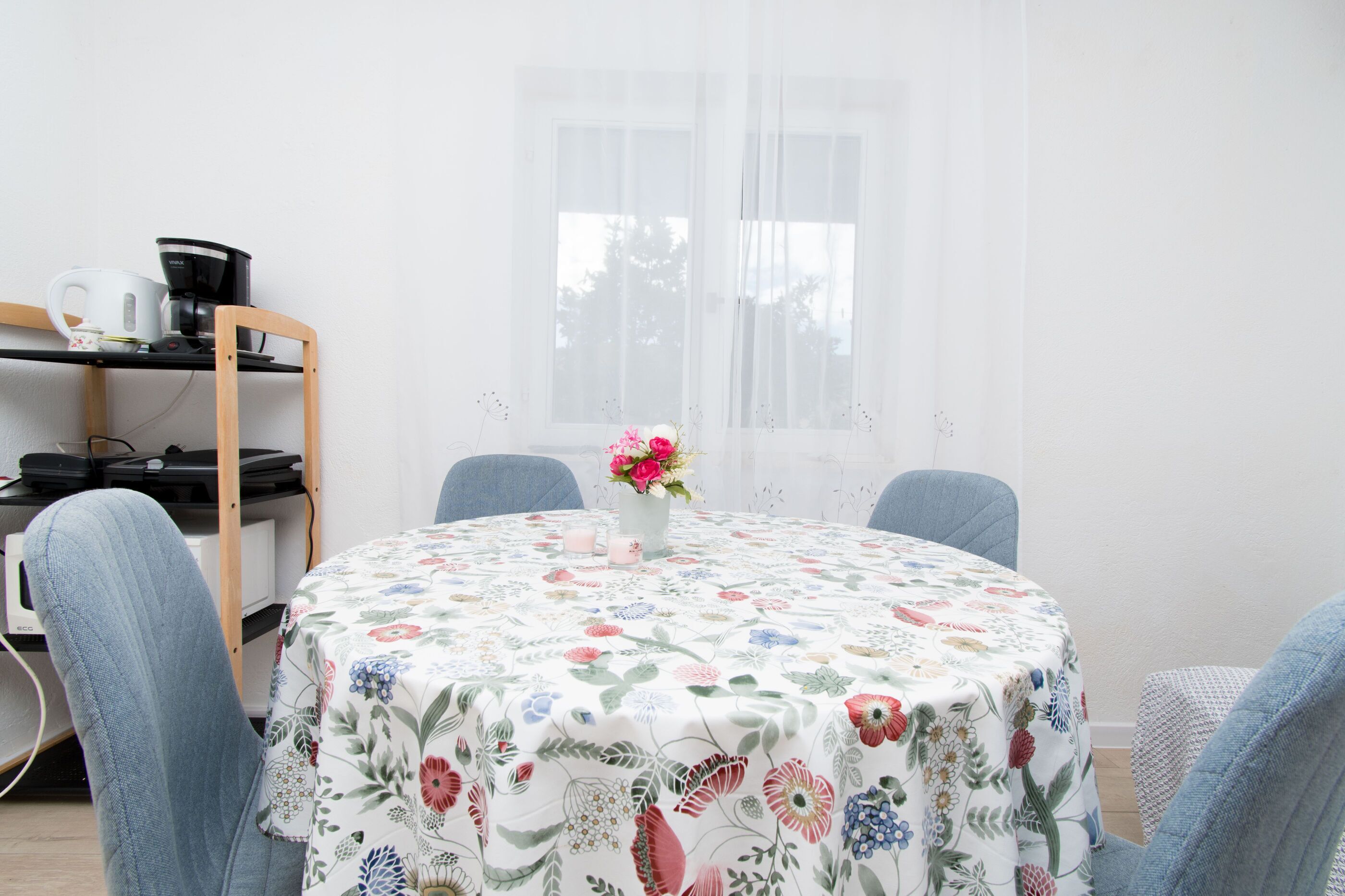 A round table with a floral tablecloth, two chairs, a vase with flowers, and a coffee maker on a shelf.