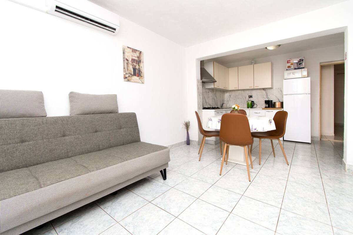 A modern living room with a sofa, dining table, chairs, and a kitchen area with cabinets and appliances.