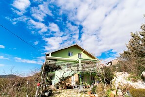 Ein zweistöckiges Haus mit grüner Fassade, einer Treppe, die zum Obergeschoss führt, und einem Gartenbereich mit Kübelpflanzen.