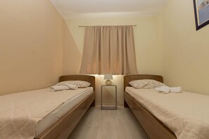 Ein kleiner, gemütlicher Raum mit zwei Einzelbetten, einem Nachttisch mit Lampe und einem Fenster mit leichtem Vorhang.