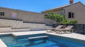 A swimming pool with lounge chairs, a stone wall, and a building with a tiled roof. - Apartments Maja (Crikvenica)