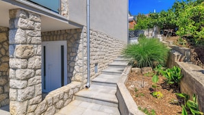 A stone exterior wall, a white door, a concrete pathway, and a garden with plants. - Apartments Maja (Crikvenica)