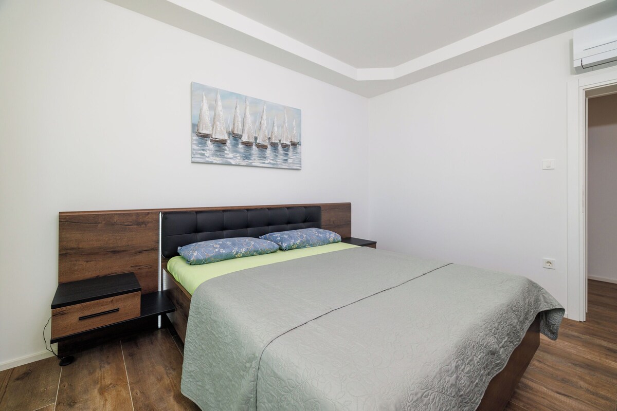 Un dormitorio con una cama, una mesita de noche, un cajón, un cuadro de veleros y una puerta.