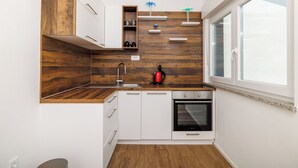 A compact kitchen with white cabinets, a wooden accent wall, a built-in oven, and a window. - Apartments Maja (Crikvenica)