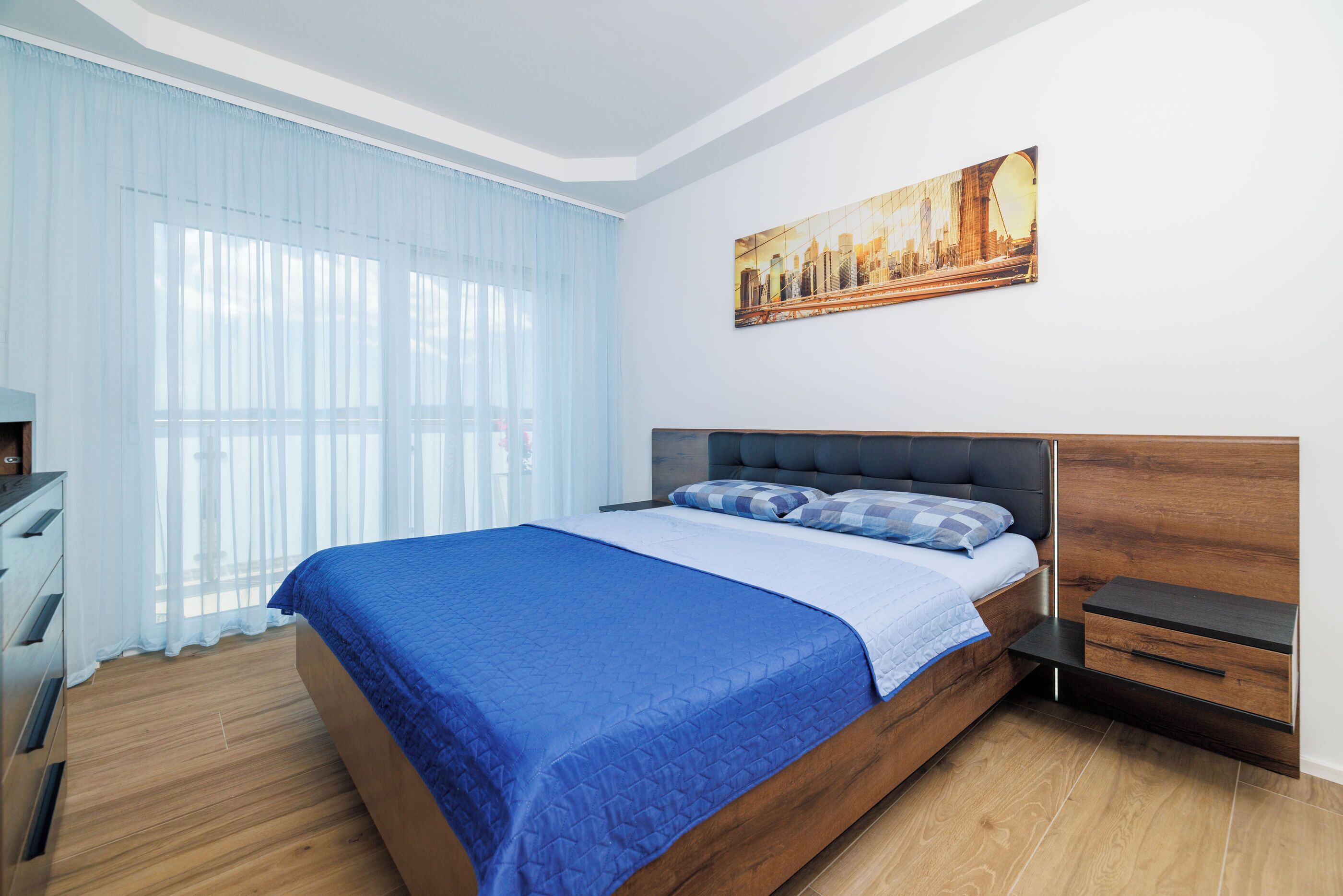 Un dormitorio con cama, mesita de noche, cómoda, ventana con cortinas translúcidas y un cuadro de paisaje urbano en la pared.