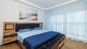A bedroom with a bed, a bench, a nightstand, a drawer, a window with curtains, and a painting on the wall. - Apartments Maja (Crikvenica)