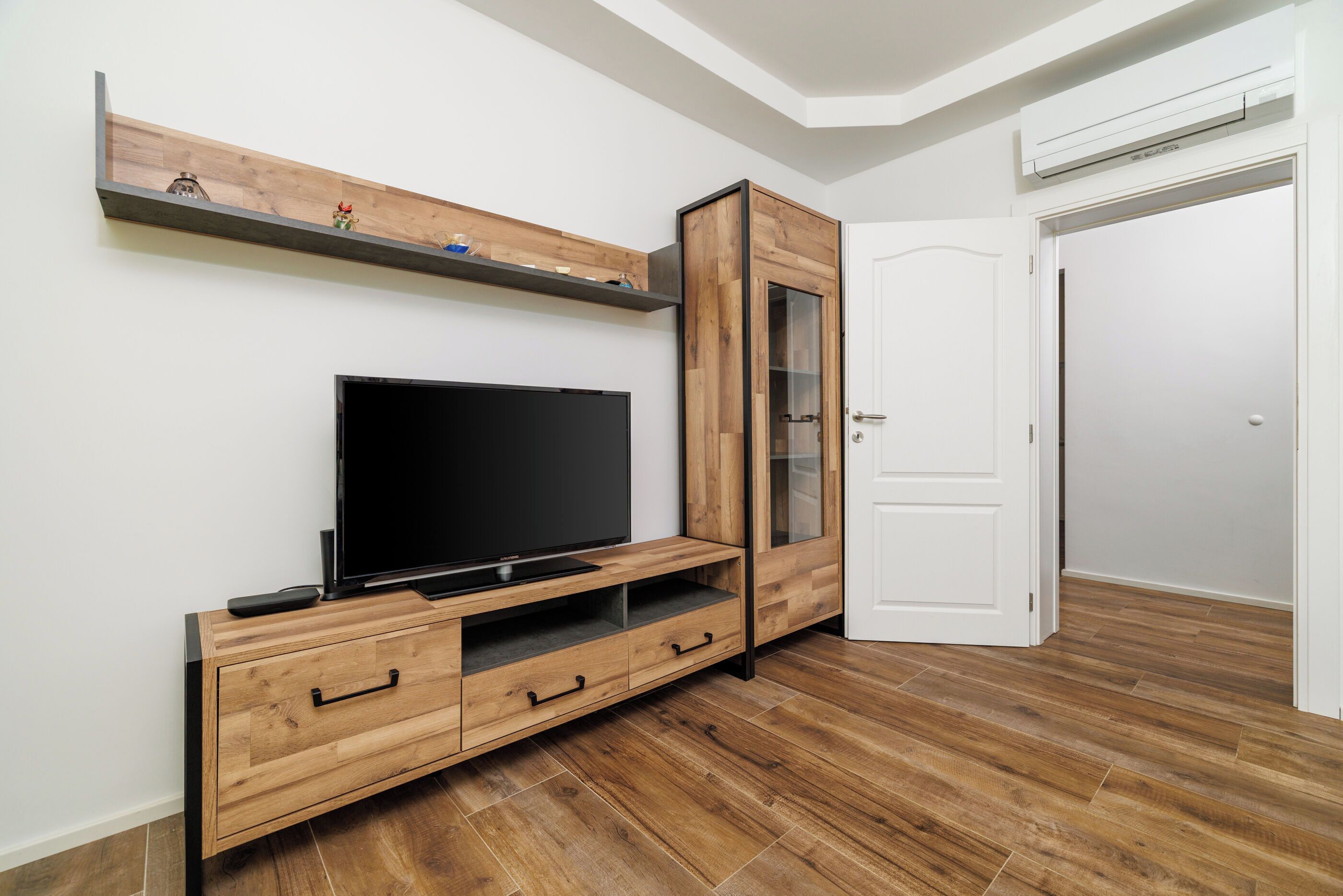 Une pièce avec un meuble de rangement en bois pour les appareils multimédias, un téléviseur à écran plat, une porte blanche et un plancher en bois.