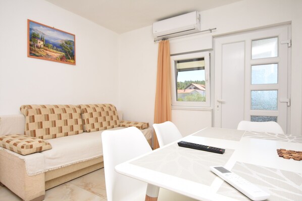 A modern room with a white sofa, a dining table with chairs, a painting on the wall, and a window with a view of houses. - Apartments Relax (Obrovac)