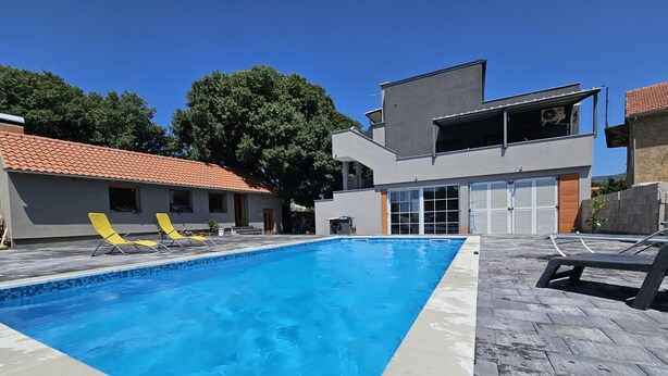 A swimming pool with lounge chairs, a modern building with a garage, and a red-tiled roof. - Apartments Relax (Obrovac)