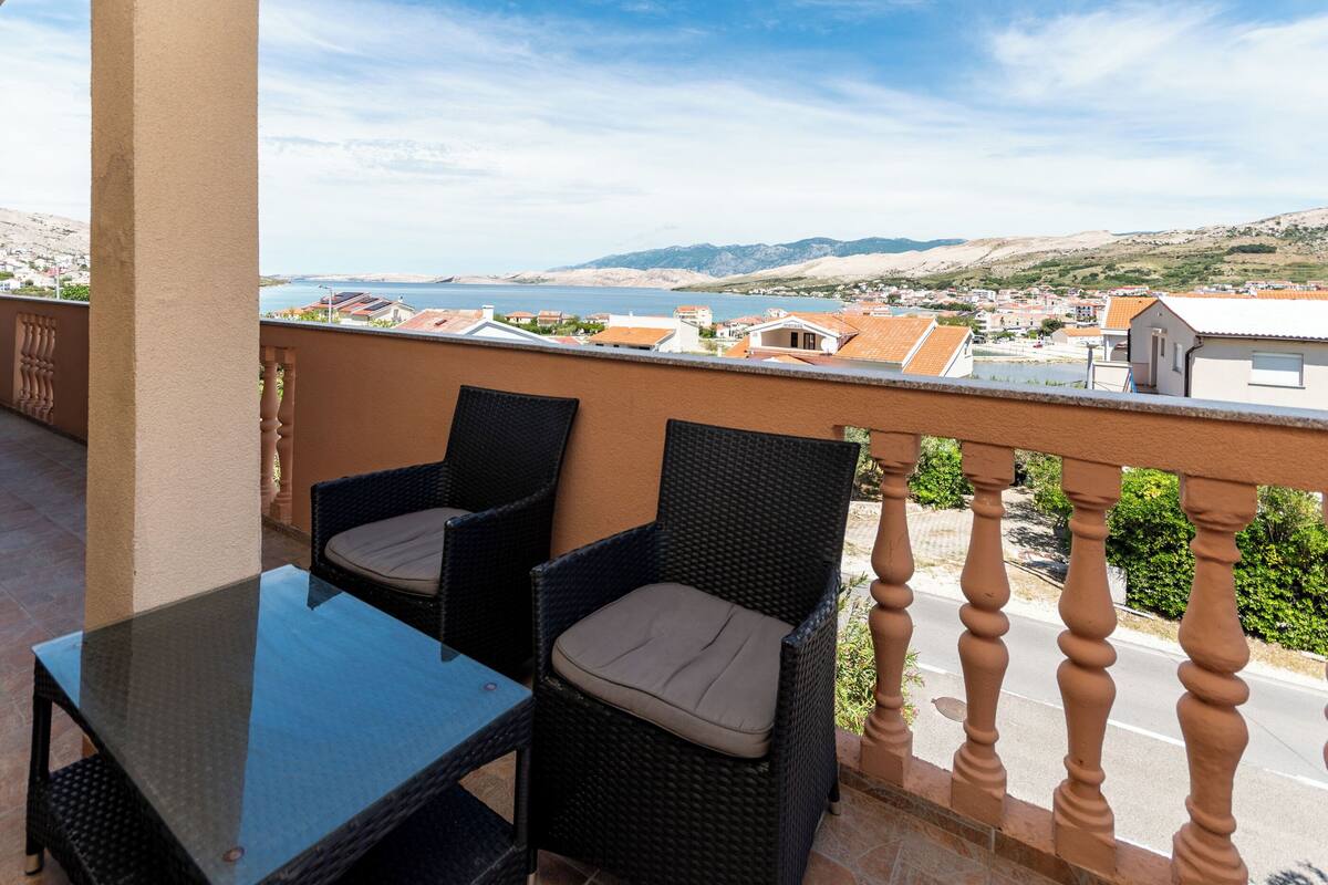 Un balcon agrémenté de fauteuils en osier et d’une table en verre, offrant une vue sur une ville côtière avec ses bâtiments et ses collines.