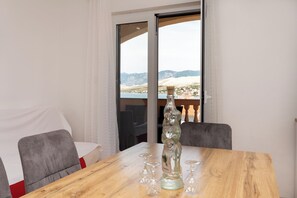 Ein Holztisch mit einer Glasflasche und Weingläsern, ein weißer Vorhang und Blick auf Berge durch eine Glastür.