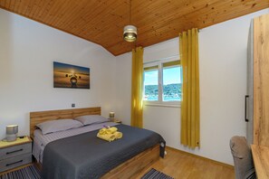 A bedroom with a wooden ceiling, a bed with a dark quilt, a painting on the wall, a window with yellow curtains, and a nightstand with a lamp. - Apartments Angela (Rab)