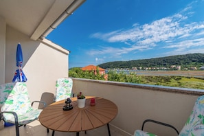 A balcony with a round wooden table, two chairs with floral upholstery, a potted plant, and a red candle. - Apartments Angela (Rab)