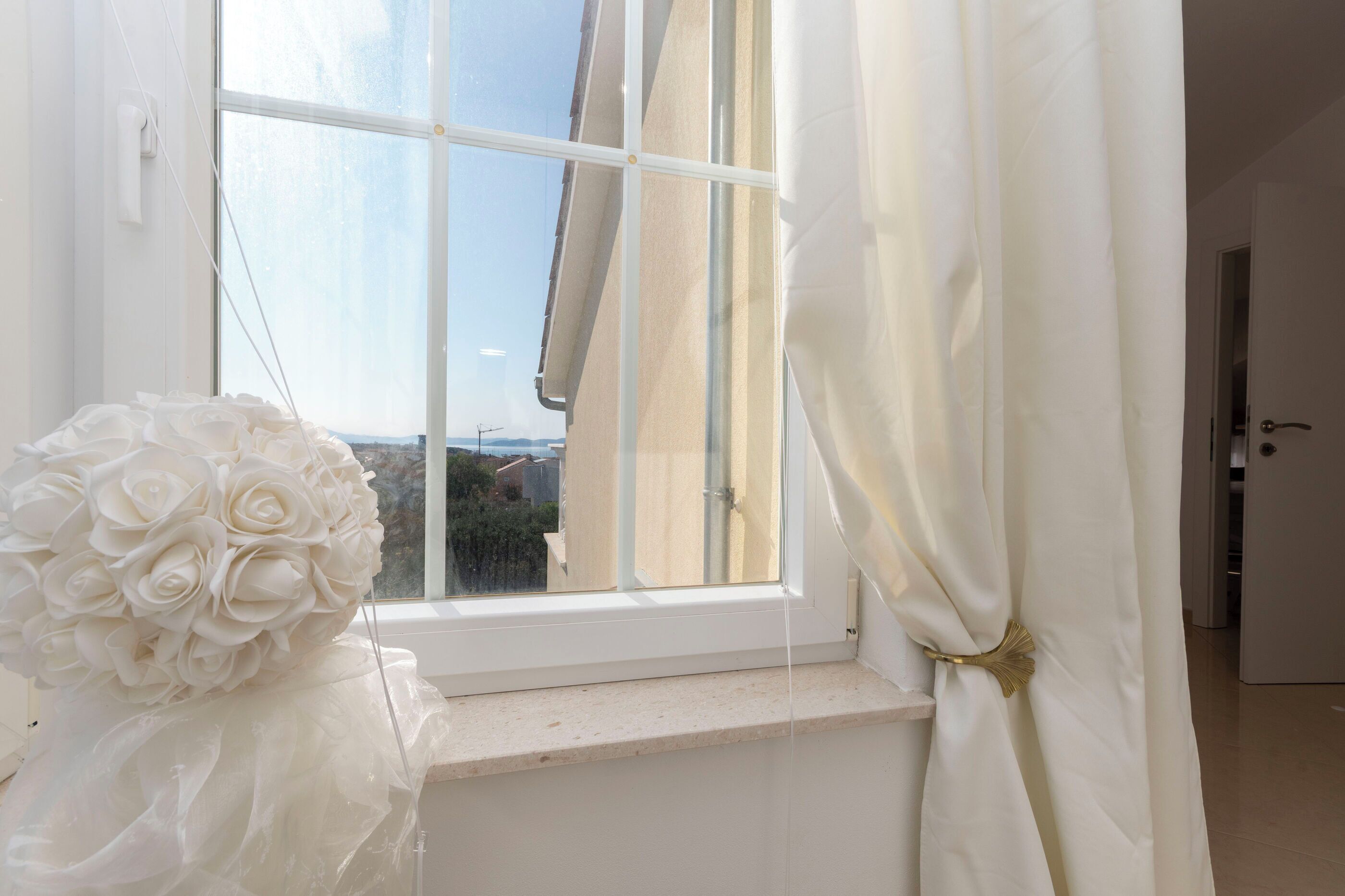 Ein Fenster mit Blick auf Gebäude, ein weißer Vorhang und ein Strauß weißer Rosen auf einem Fensterbrett.