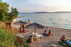Ein Kiesstrand mit Sonnenschirmen, Liegestühlen und Menschen, die am Wasser entspannen.