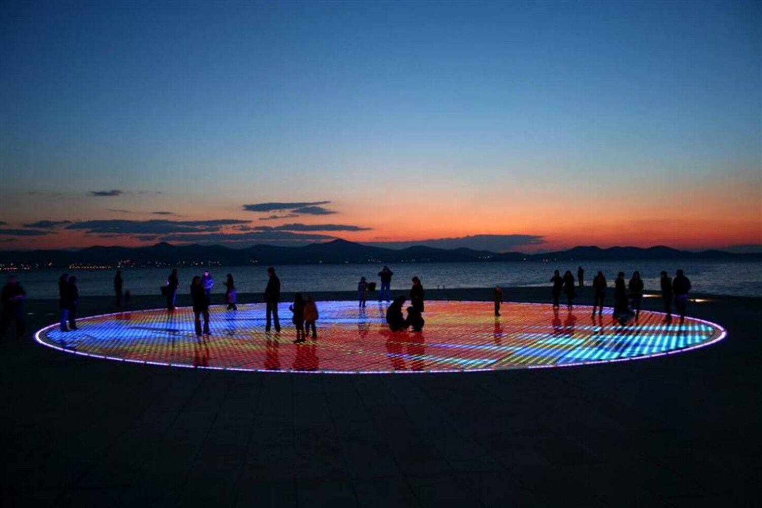 A circular LED-lit platform with people around it, overlooking a body of water at dusk.