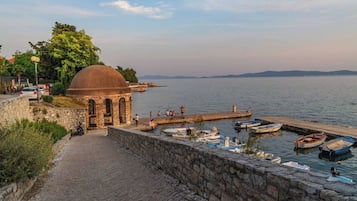 Uma trilha de pedra leva a um edifício histórico com uma cúpula, uma orla com barcos e uma vista clara das montanhas ao longe.