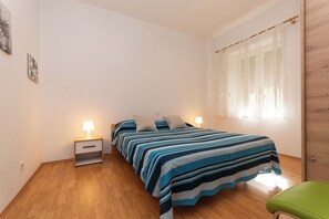 A bedroom with a bed, nightstands, a green chair, and a window with sheer curtains. - Apartment Kalma (Zadar)