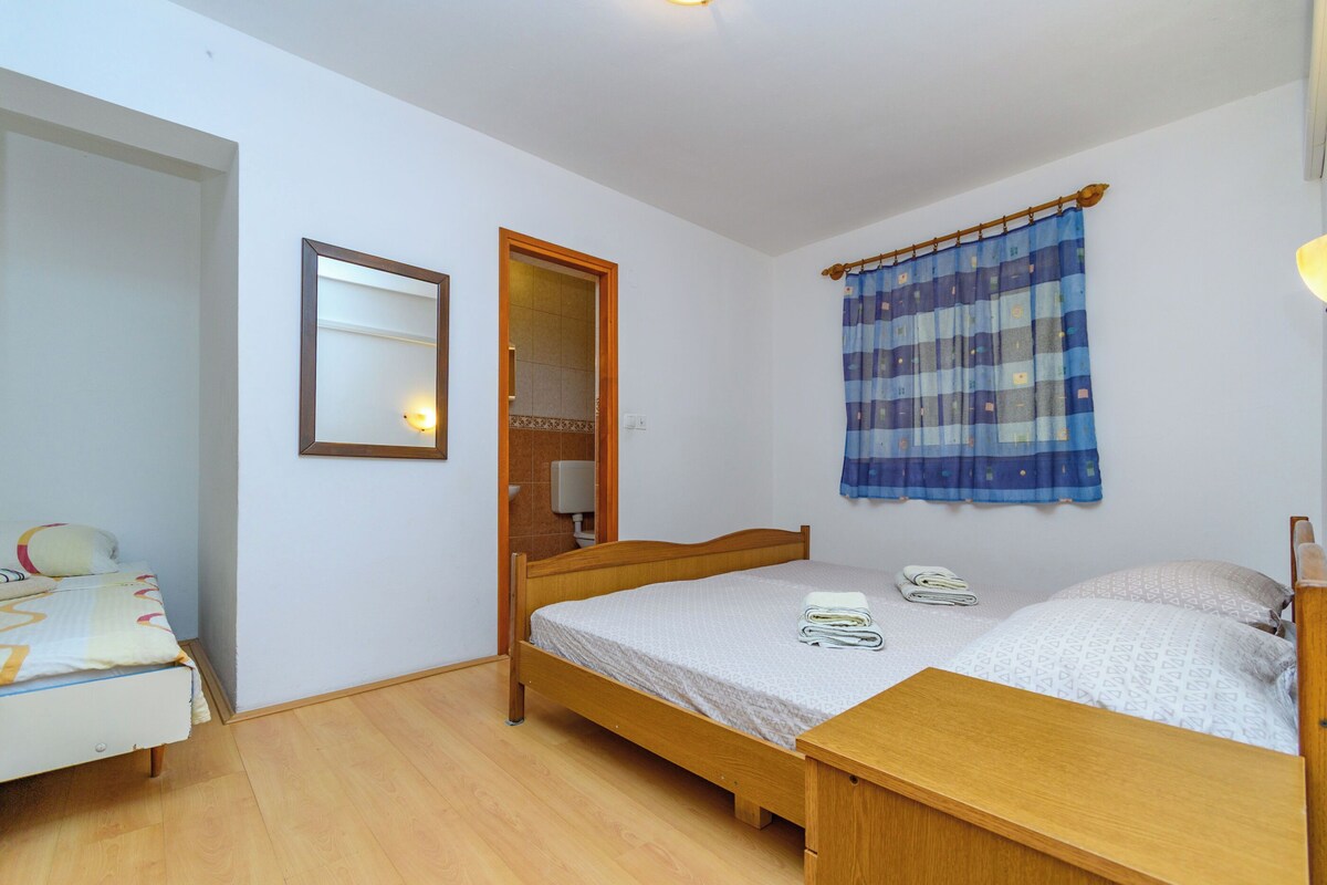 Ein kleines, ordentliches Schlafzimmer mit einem Einzelbett, einem Nachttisch aus Holz, einem Spiegel und einem Fenster mit blauer Gardine.