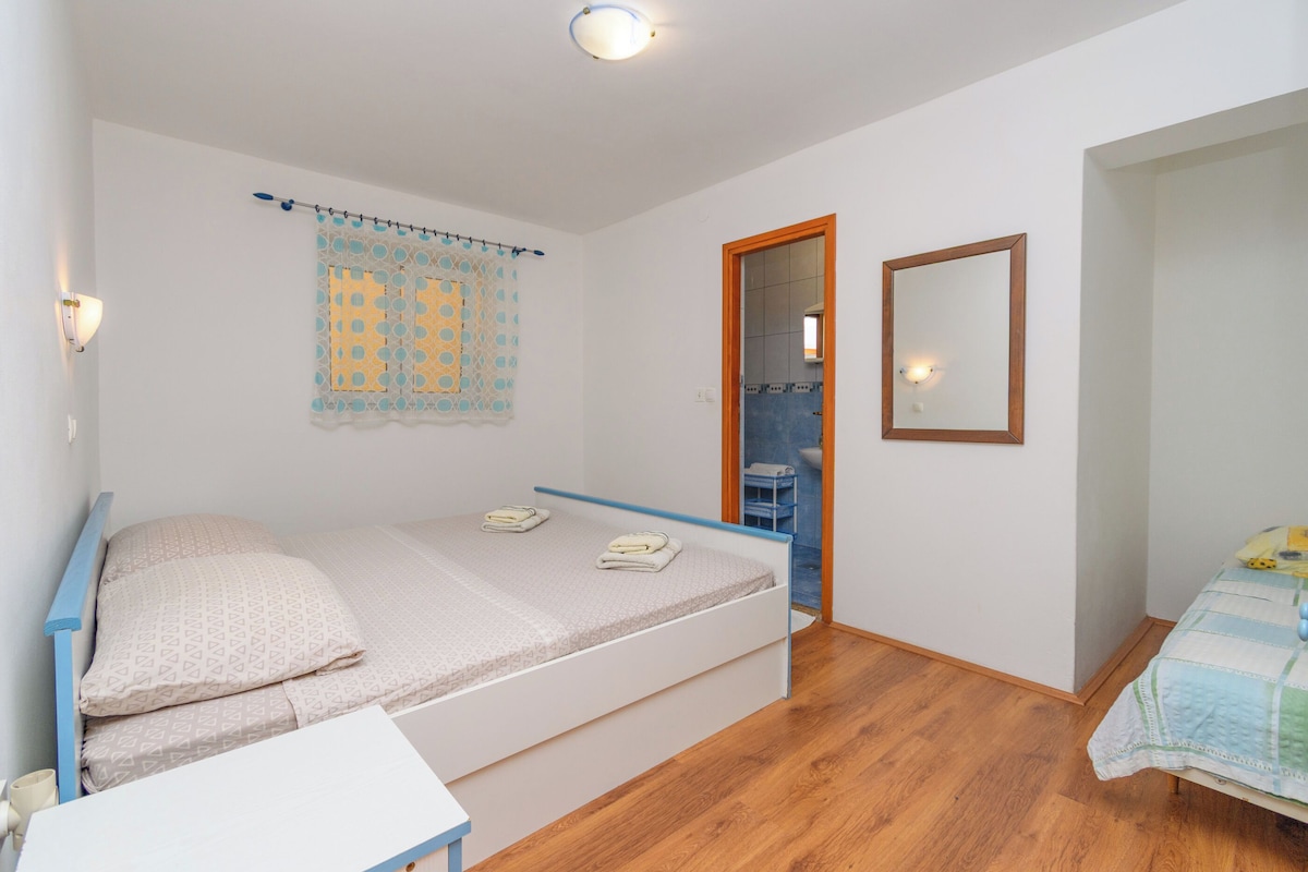 Einzimmerappartement mit Holzboden, einem kleinen Tisch, einer Wandleuchte, einem Fenster mit Vorhang und einer Tür zu einem Badezimmer.