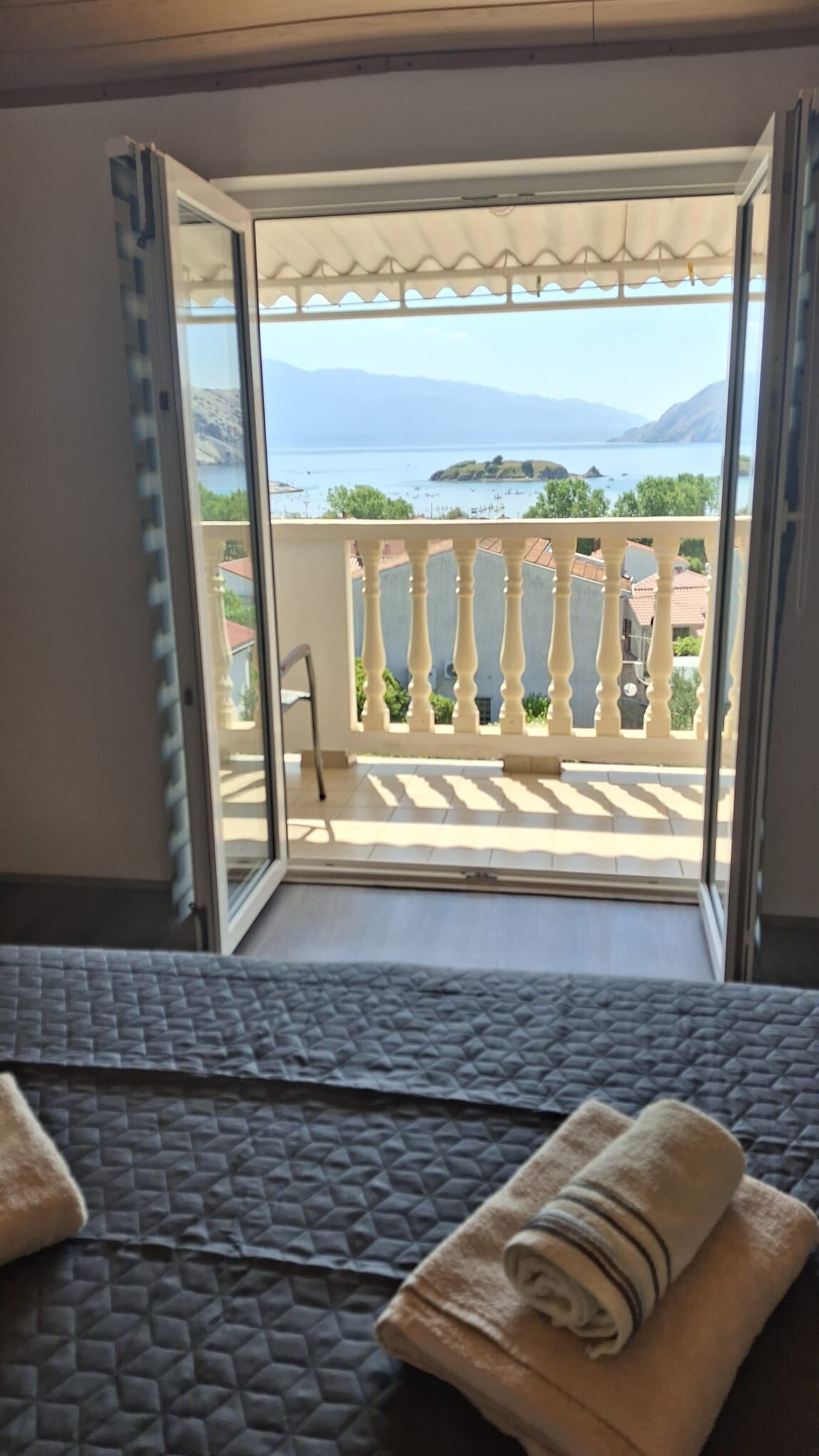 A room with a bed, a view of a balcony with a railing, and a glimpse of the sea and mountains in the distance.