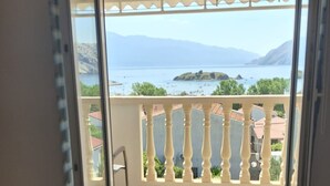 Quarto com uma cama, vista para uma varanda com guarda-corpo e um vislumbre do mar e montanhas ao longe.