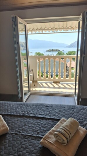 Ein Zimmer mit einem Bett, Blick auf einen Balkon mit Geländer und einen Ausblick auf Meer und Berge in der Ferne.