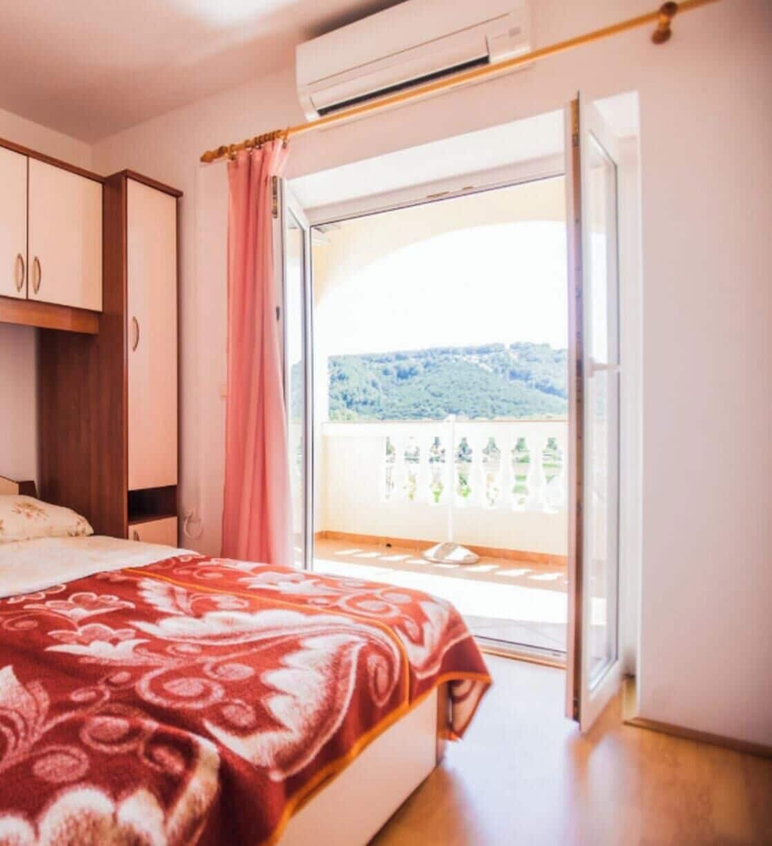 Ein Schlafzimmer mit Bett, Schrank und einem Balkon mit Blick auf Grünflächen und Hügel.