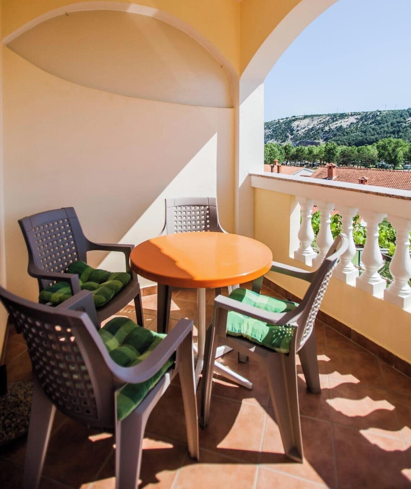 Ein Balkon mit einem runden Tisch und vier Stühlen, jeder mit grünen Kissen. Der Balkon verfügt über eine weiße Brüstung und bietet Blick auf eine hügelige Landschaft mit Bäumen und Gebäuden in der Ferne.