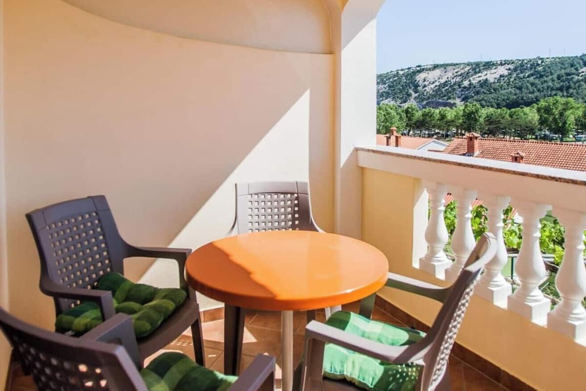 A balcony with a round table and four chairs, each with green cushions. The balcony has a white balustrade and offers a view of a hilly landscape with trees and buildings in the distance.