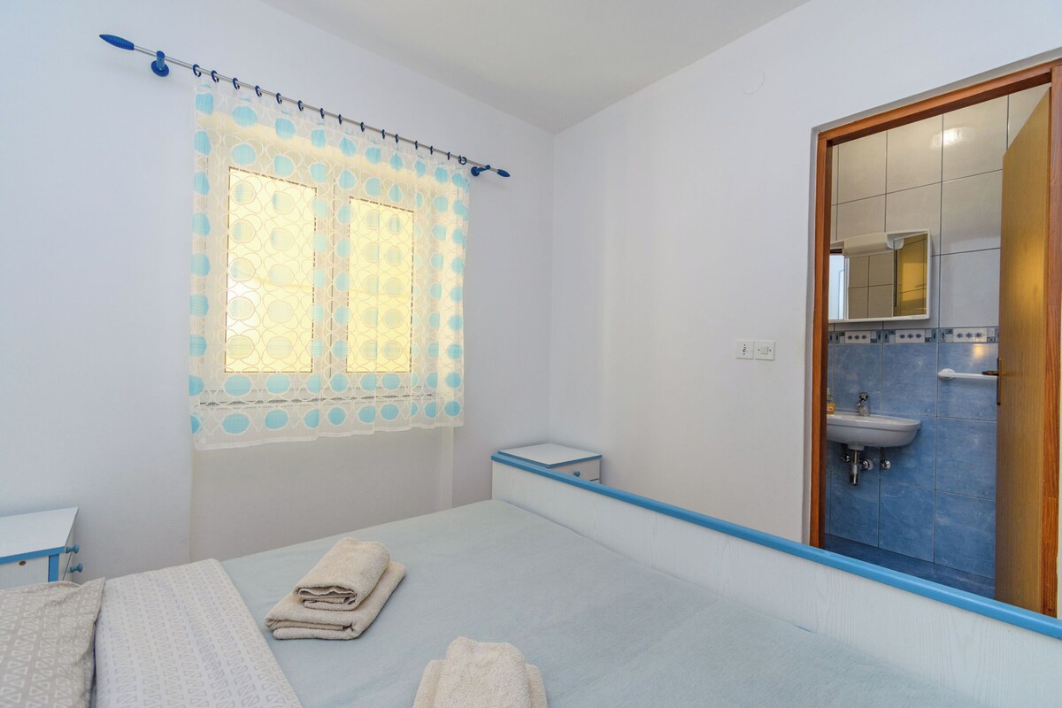 Ein Schlafzimmer mit Bett, Fenster mit Vorhang, Nachttisch und einem Badezimmer mit Waschbecken und Spiegel.