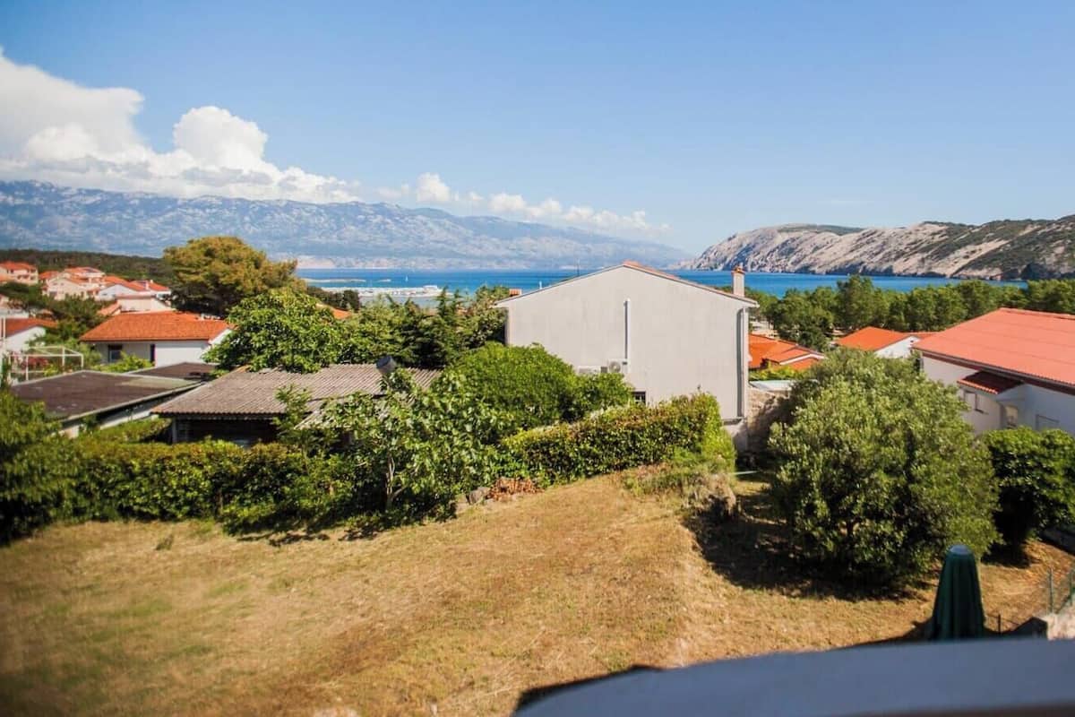 A coastal village with houses, a clear blue sea, and mountains in the background.
