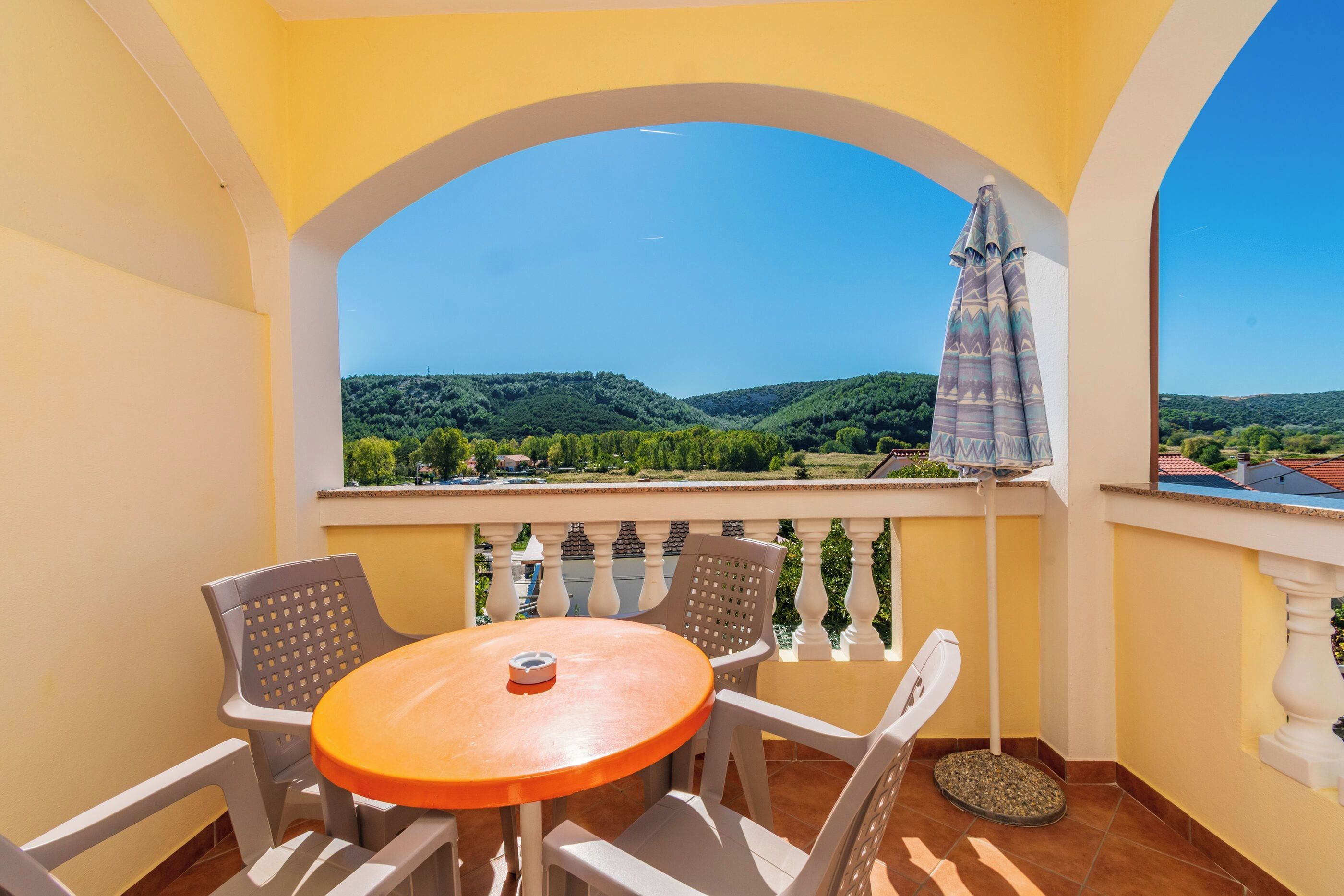 A balcony with a table and chairs, a view of a green landscape, and a patterned umbrella.