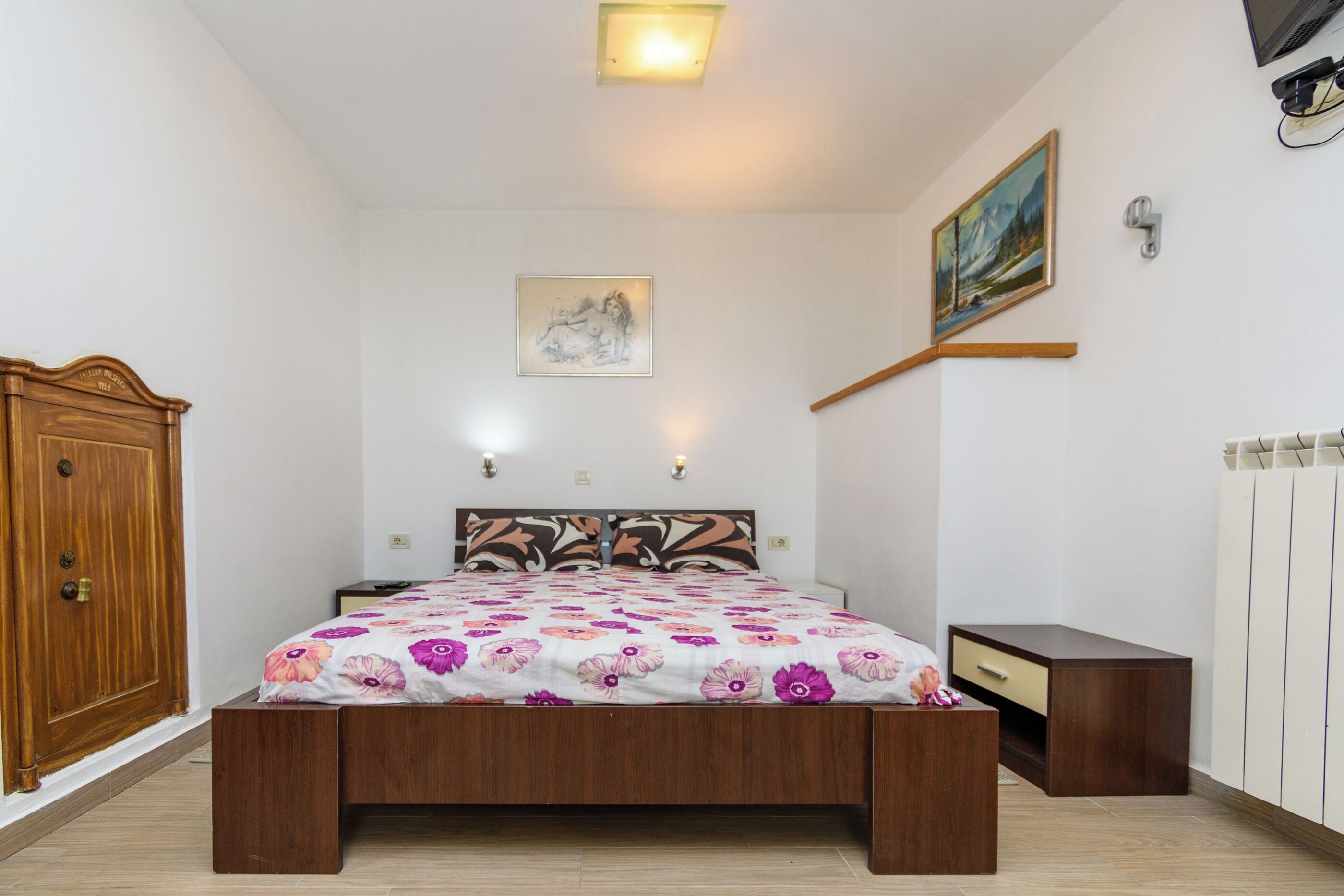 Ein Schlafzimmer mit einem hölzernen Bett, einem Nachttisch, einem Kleiderschrank, einem Gemälde und einer Heizung.