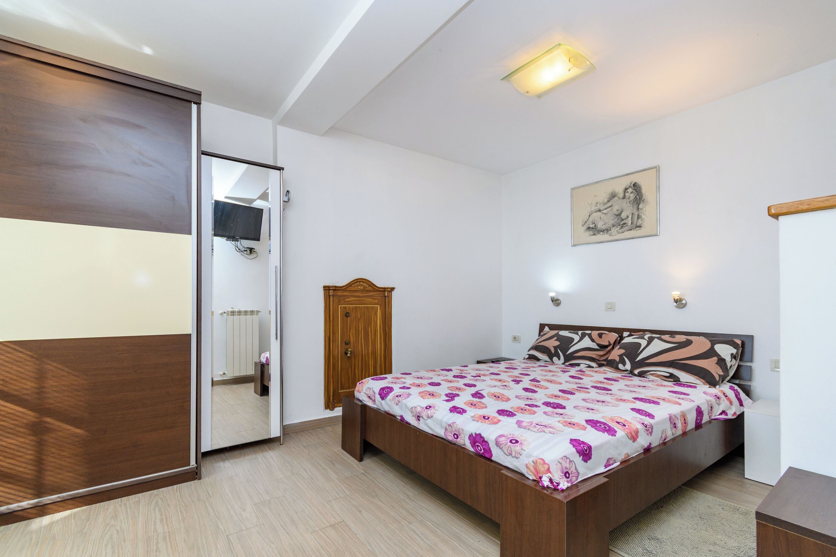 A bedroom with a bed, a wooden wardrobe, a door, a wall-mounted light, and a painting.