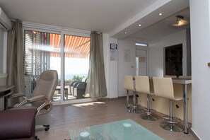 Una habitación con puerta de vidrio corrediza que da a un balcón, un área de descanso con sillas, una barra con taburetes y una kitchenette visible a través de la puerta.