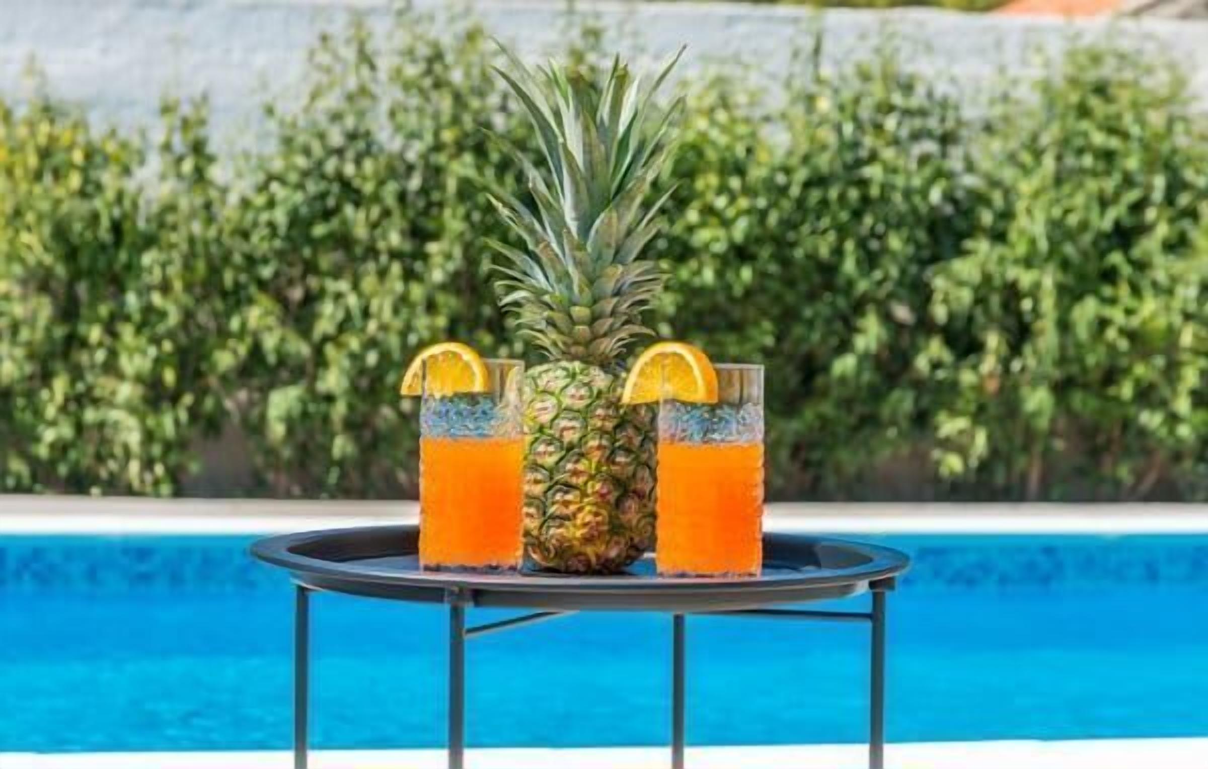 Una mesa con dos vasos de una bebida de piña y naranja, una piña y una piscina al fondo.