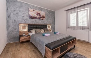 Ein Schlafzimmer mit einem Bett, Nachttischen, einer Bank, einem Gemälde und einem Fenster mit Vorhängen.