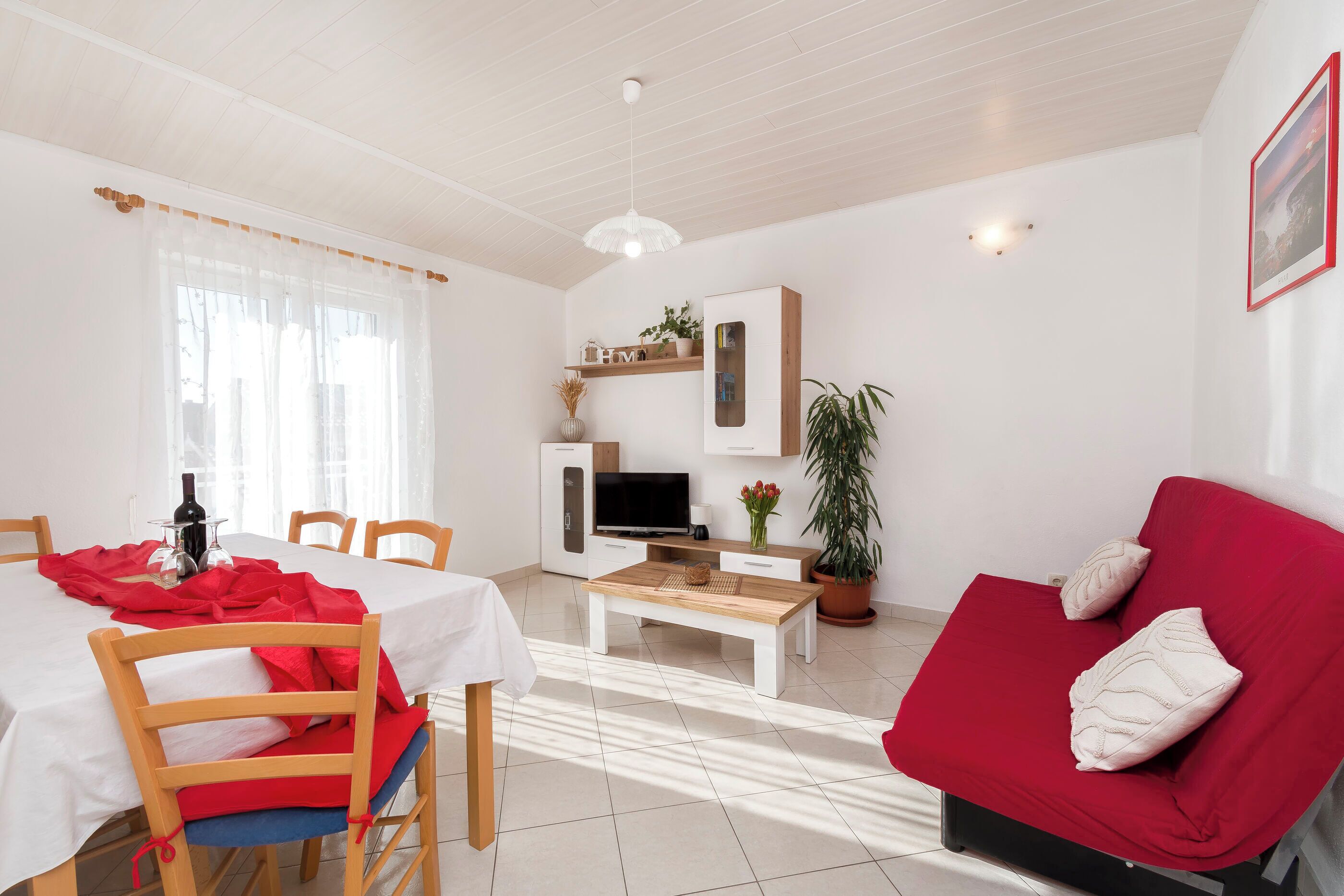 A room with a dining table, chairs, a red sofa, a TV, and a potted plant.
