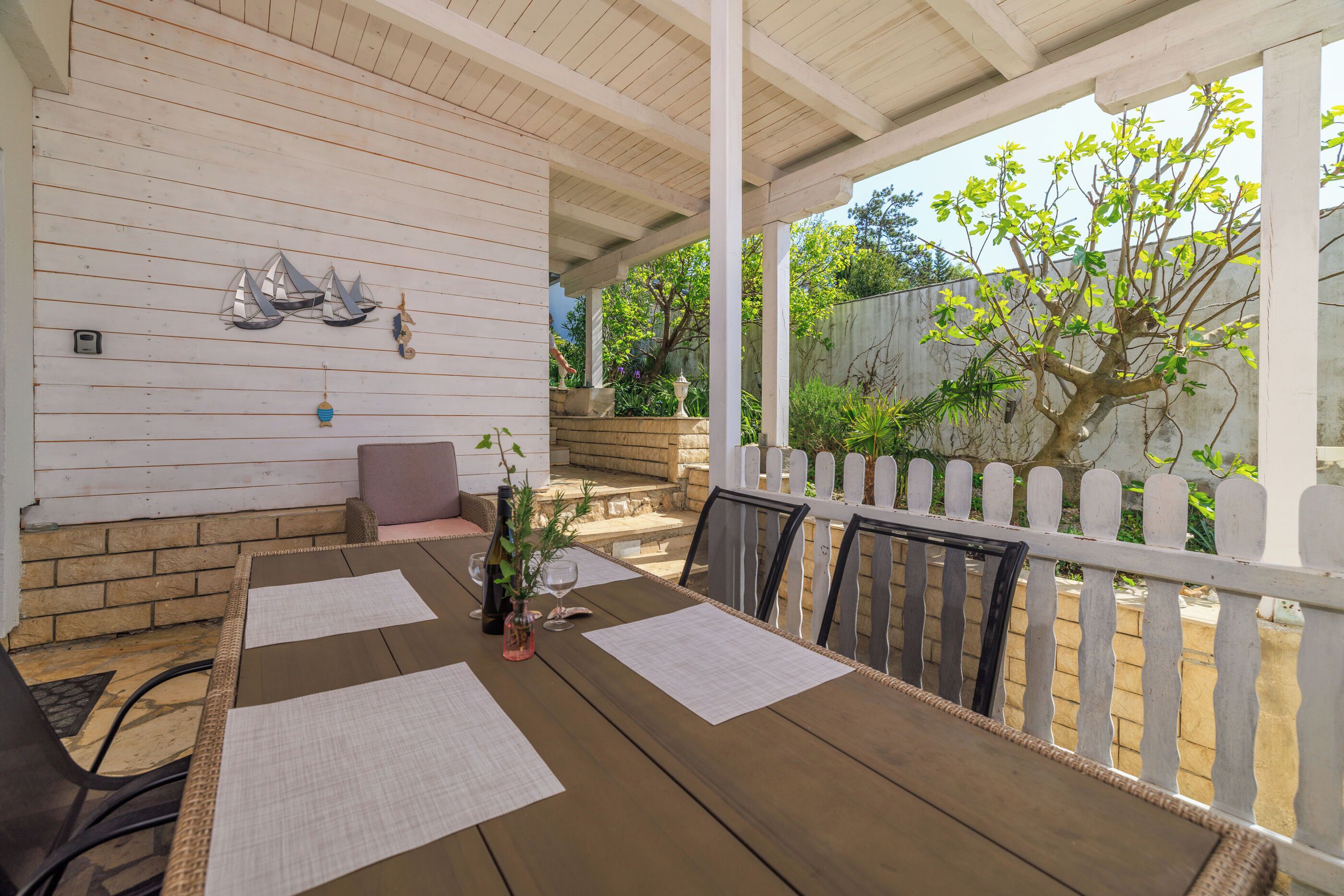 Une terrasse couverte avec une table en bois dressée pour les repas, des rampes blanches et une vue sur la verdure.
