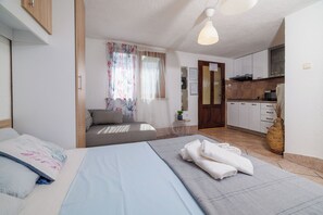 Ein Schlafzimmer mit Bett, Nachttisch, einem Fenster mit Vorhängen, einer kleinen Küchenzeile mit Schränken und einem Herd sowie einer Tür zu einem anderen Raum.
