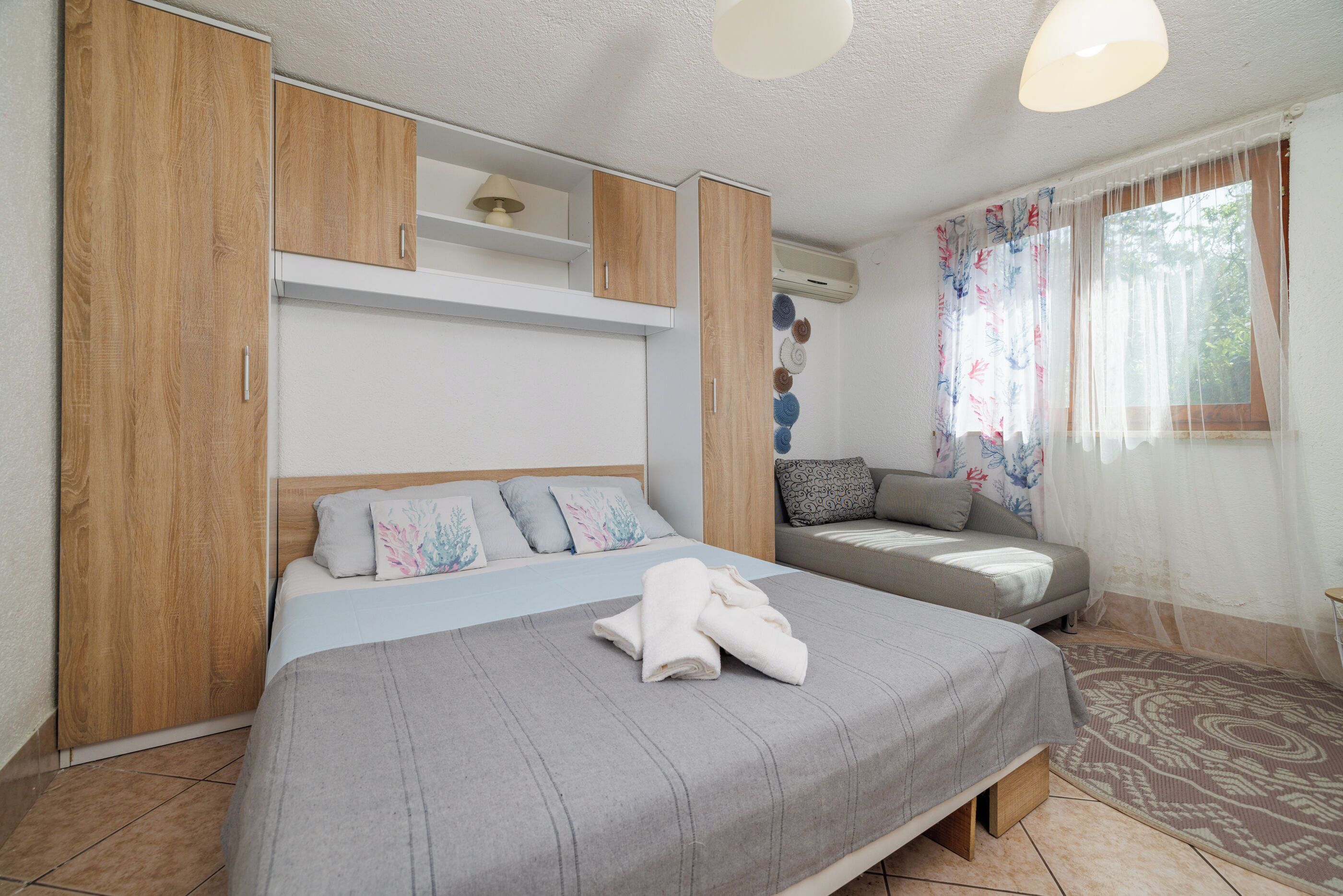 Ein Schlafzimmer mit einem Bett, einer Couch, Holzschränken, einer Deckenlampe und einem Fenster mit Vorhängen.