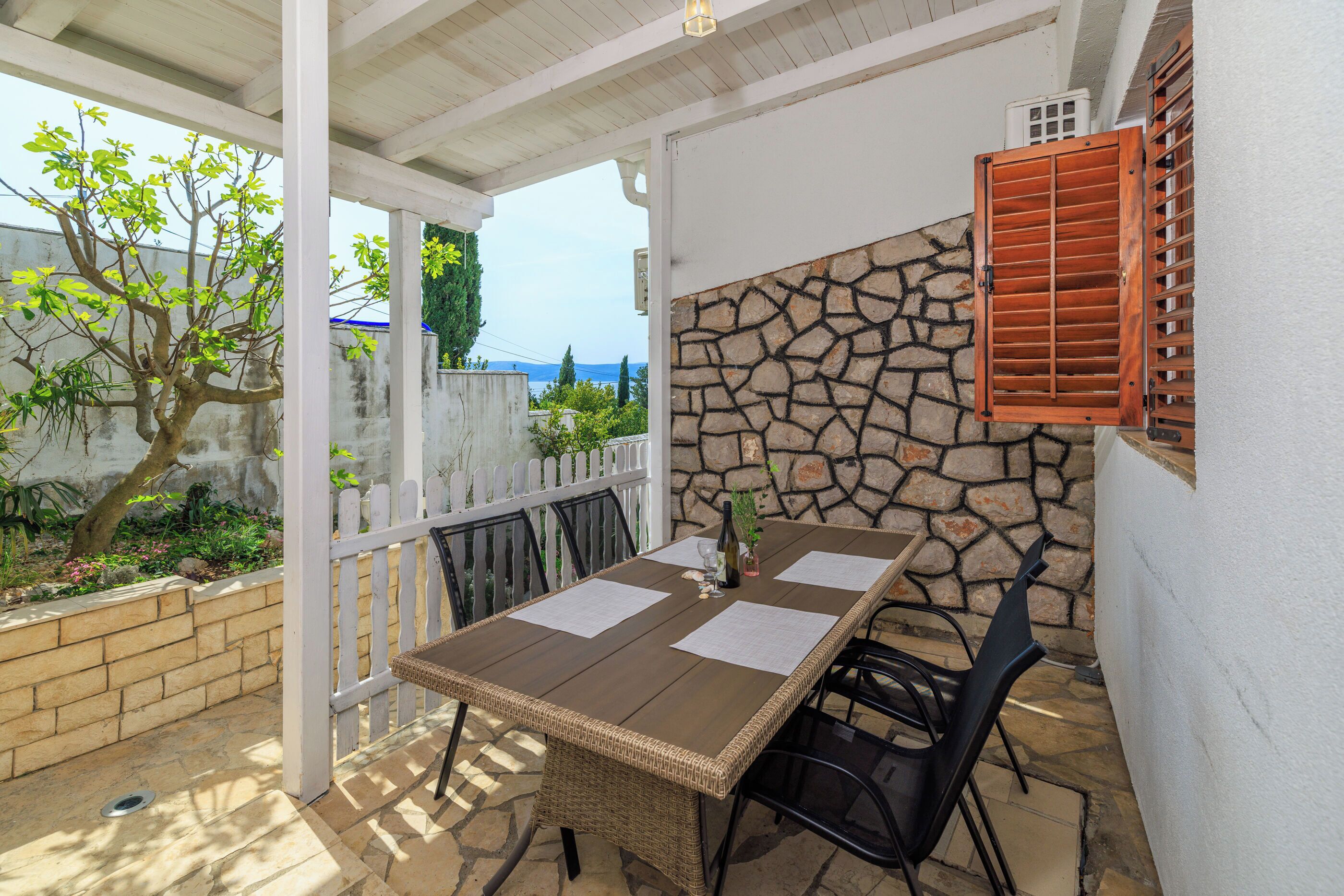 An outdoor patio with a table and chairs, a stone wall, and a view of greenery and a building in the background.