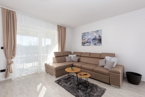 Ein modernes Wohnzimmer mit einem Ecksofa, zwei kleinen Tischen, einem schwarzen Teppich, einem Gemälde an der Wand und durchsichtigen Vorhängen.
